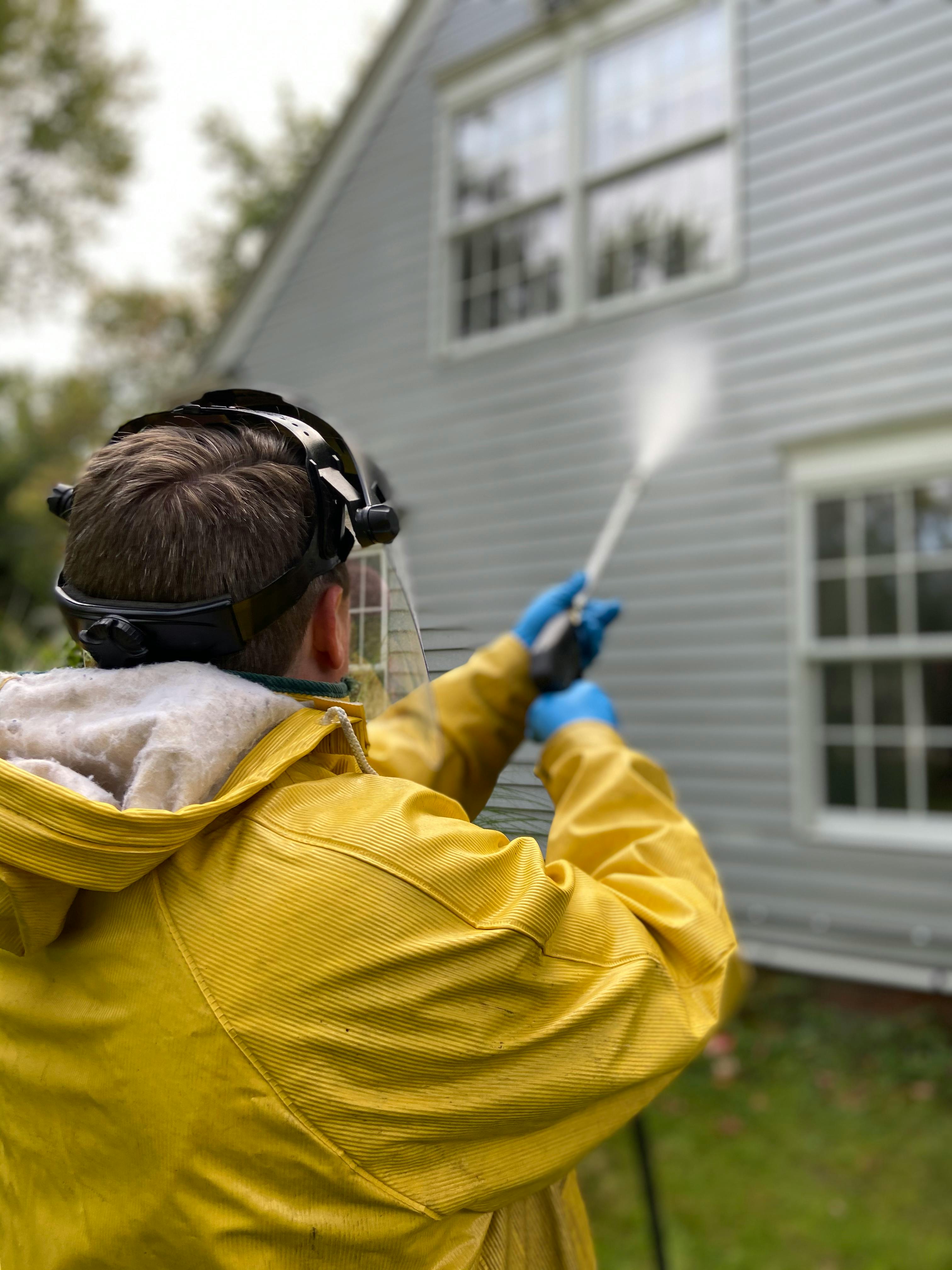 Worker in yellow safety gear pressure washing house exterior siding - professional residential cleaning service removes dirt, mold, and weather stains from home walls